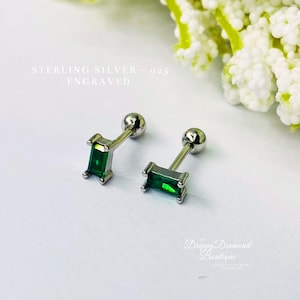 May include: Two sterling silver stud earrings with emerald green rectangular stones. The earrings have a post and ball backing. The text "STERLING SILVER - 925 ENGRAVED" is visible. The earrings are displayed on a white surface.