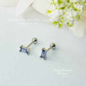 May include: A pair of sterling silver 925 earrings with a small, rectangular, light blue gemstone. The earrings are designed for cartilage piercings.