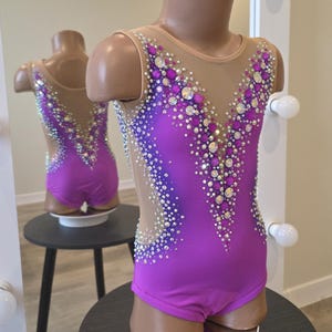 Peut inclure: Un justaucorps sans manches en tissu violet vif avec un d&eacute;collet&eacute; en V profond et des empi&egrave;cements lat&eacute;raux en maille nude. Le v&ecirc;tement est richement orn&eacute; de cristaux transparents et violets dispos&eacute;s en un motif dense et scintillant.