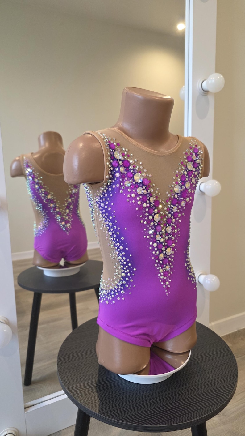 Peut inclure: Un justaucorps sans manches en tissu violet vif avec un d&eacute;collet&eacute; en V profond et des empi&egrave;cements lat&eacute;raux en maille nude. Le v&ecirc;tement est richement orn&eacute; de cristaux transparents et violets dispos&eacute;s en un motif dense et scintillant.
