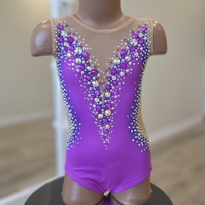 Peut inclure: Un justaucorps sans manches d'un violet vif dot&eacute; d'un d&eacute;collet&eacute; en V profond avec un tissu en maille beige transparent. Le v&ecirc;tement est orn&eacute; de nombreux cristaux transparents et violets dispos&eacute;s en un motif dense et &eacute;tincelant.