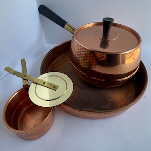 May include: A collection of copper cookware including a large round tray, a smaller copper pot with a black handle and lid, and a small copper cup with a brass lid featuring a cross-shaped handle mechanism. The items have a polished finish.
