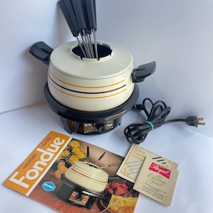 May include: An off-white electric fondue pot with two brown horizontal stripes, black handles, and a black base. Six black-handled fondue forks are inserted into the pot. Includes an instruction manual titled automatic fondue and a Regal brand card.