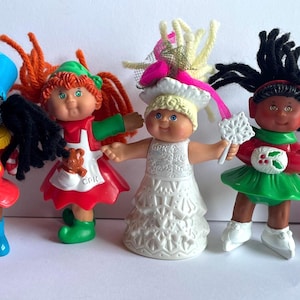 May include: Five small plastic dolls with yarn hair, each wearing different outfits: a blue and red soldier uniform, a red and green holiday dress, a white lace gown with a pink bow, a green dress with holly, and a pink dress with a yellow bear.