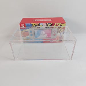 May include: A clear acrylic display case with a Nintendo Switch Lite console and a Pokemon game box inside. The console is white with blue and pink accents. The game box is red with a Pokemon image on the front.