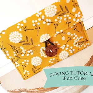 May include: A yellow fabric iPad case with a white floral pattern and a brown button closure. The case is sitting on a white surface with the text "Sewing Tutorial iPad Case" on a blue background.