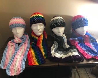 May include: Four crocheted hats and scarves in various colours, including pink, blue, white, grey, black, and rainbow. The hats and scarves are displayed on a wooden surface.