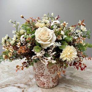 May include: A floral arrangement featuring a large cream rose and a pale yellow dahlia surrounded by small white flowers, green foliage, and rust-colored sprigs. The flowers are contained in a cylindrical pot with a brown floral pattern.