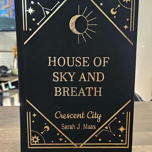 Puede incluir: Un libro de tapa dura negro con letras e ilustraciones doradas. El título es "House of Sky and Breath" y el subtítulo es "Crescent City" de Sarah J. Maas.