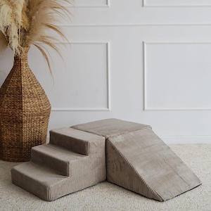 Può includere: Un set da gioco beige per bambini, con rampa e gradini in velluto a coste. Sono presenti vasi intrecciati con piante essiccate. Lo sfondo è una parete bianca con pannelli. Il testo "Hand Made in Poland" è visibile.