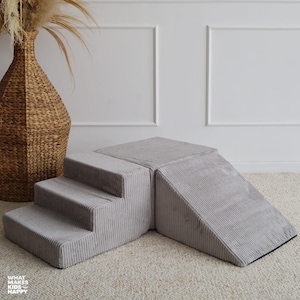 Può includere: Un set da gioco in schiuma di velluto a coste grigio chiaro con gradini e una rampa. Il set è progettato per i bambini ed è posto su un tappeto neutro. Un vaso intrecciato con piante essiccate è sullo sfondo.