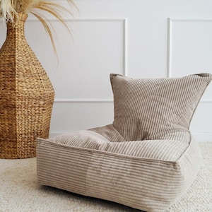 May include: A beige corduroy beanbag chair with a high back and a woven vase filled with dried plants. A white rocking horse is on the left. The room has white walls and a light-coloured rug.