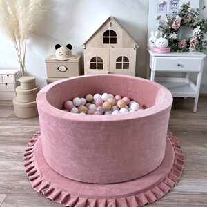May include: A pink velvet ball pit with a ruffled base. The pit is filled with colorful balls and is surrounded by a wooden dollhouse, a white nightstand, and a white board with the text "Enjoy the little things".