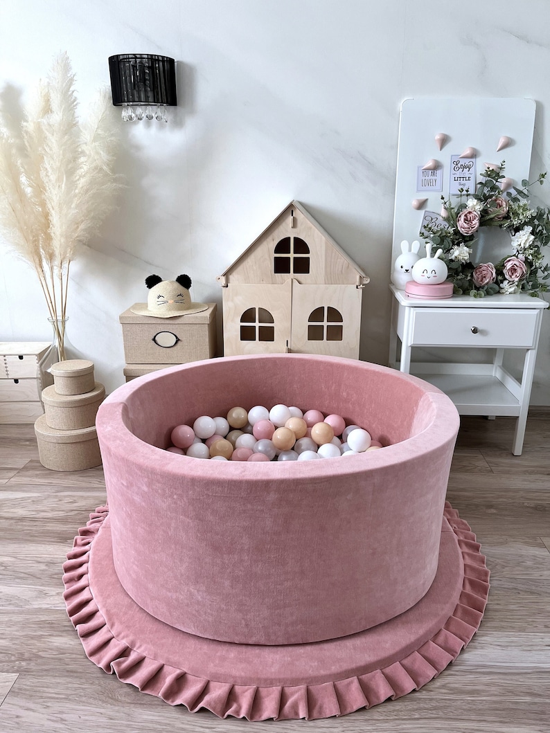 May include: A pink velvet ball pit with a ruffled base. The pit is filled with colorful balls and is surrounded by a wooden dollhouse, a white nightstand, and a white board with the text "Enjoy the little things".