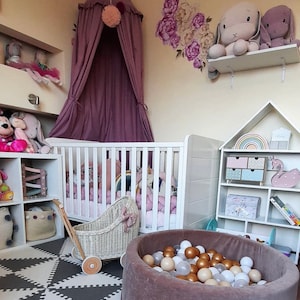 Puede incluir: Una cuna blanca con un juego de ropa de cama rosa y blanca, un cochecito de mu&ntilde;ecas de mimbre blanco, una piscina de bolas marr&oacute;n llena de bolas blancas y doradas, y una estanter&iacute;a blanca con un arco&iacute;ris y una figurita de cisne rosa. La habitaci&oacute;n est&aacute; decorada con un dosel morado, una calcoman&iacute;a de pared floral y animales de peluche.