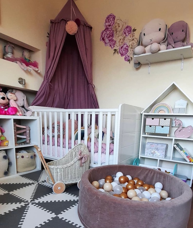 Puede incluir: Una cuna blanca con un juego de ropa de cama rosa y blanca, un cochecito de mu&ntilde;ecas de mimbre blanco, una piscina de bolas marr&oacute;n llena de bolas blancas y doradas, y una estanter&iacute;a blanca con un arco&iacute;ris y una figurita de cisne rosa. La habitaci&oacute;n est&aacute; decorada con un dosel morado, una calcoman&iacute;a de pared floral y animales de peluche.