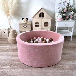 May include: A pink velvet ball pit filled with white and brown balls. The pit is round and has a soft, plush texture.