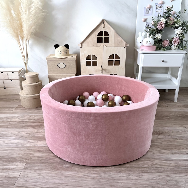 May include: A pink velvet ball pit filled with white and brown balls. The pit is round and has a soft, plush texture.