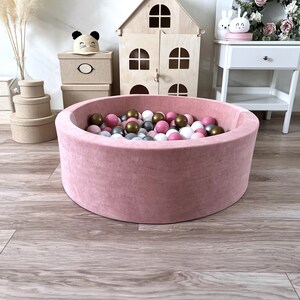 May include: A pink velvet ball pit filled with pink, white, and gold balls. The pit is round and has a soft, plush texture.