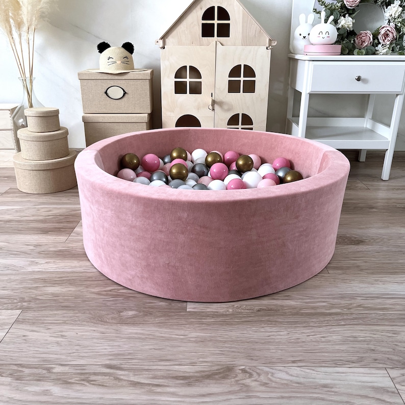 May include: A pink velvet ball pit filled with pink, white, and gold balls. The pit is round and has a soft, plush texture.