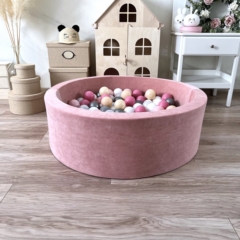 May include: A pink velvet ball pit filled with colorful balls. The pit is round and has a soft, plush texture. The balls are pink, white, and gray.