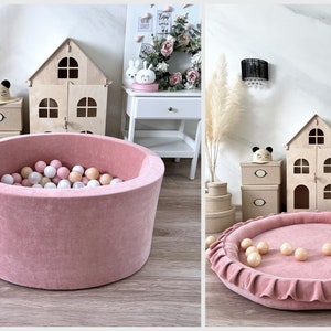 May include: A pink velvet ball pit with a ruffled edge and white and pink balls inside. The pit is sitting on a wooden floor.