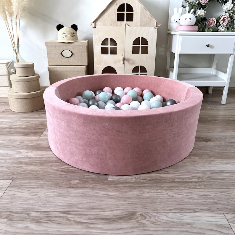 May include: A pink velvet ball pit filled with colorful balls. The pit is round and has a soft, plush texture.