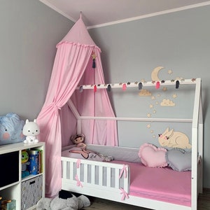 Puede incluir: Una cama de madera blanca con forma de casa con dosel rosa, ropa de cama rosa y un colch&oacute;n a rayas gris y blanco. La cama tiene una almohada en forma de coraz&oacute;n y un recorte de conejo de madera en la cabecera. La habitaci&oacute;n est&aacute; decorada con estrellas y nubes de madera.