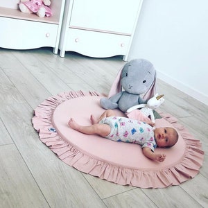 Puede incluir: Una alfombra de juego rosa con un borde de volantes, con un beb&eacute; acostado en la alfombra con un conejito gris y un juguete de peluche de unicornio.