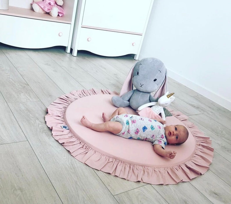 Puede incluir: Una alfombra de juego rosa con un borde de volantes, con un beb&eacute; acostado en la alfombra con un conejito gris y un juguete de peluche de unicornio.