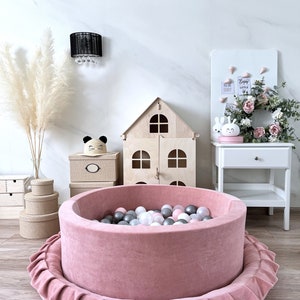 May include: A pink velvet ball pit with a ruffled edge, filled with colorful balls. The pit is in a room with a wooden dollhouse, a white nightstand, and a white board with the text "You are loved" and "Enjoy the little things".