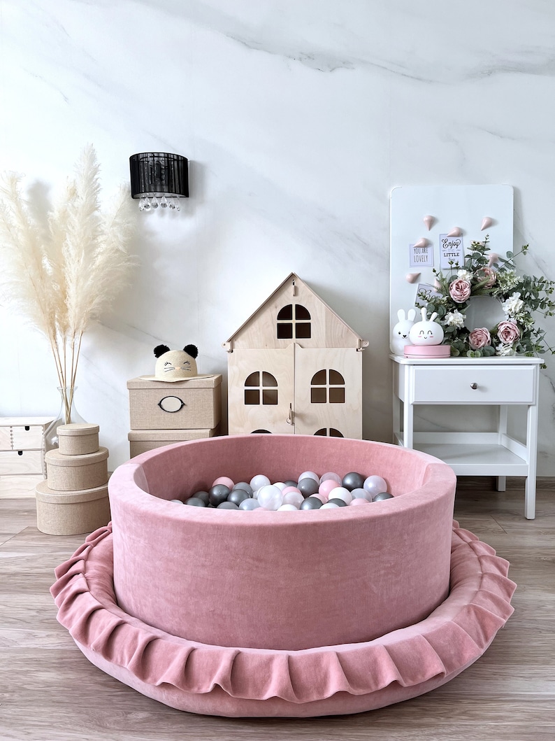 May include: A pink velvet ball pit with a ruffled edge, filled with colorful balls. The pit is in a room with a wooden dollhouse, a white nightstand, and a white board with the text "You are loved" and "Enjoy the little things".