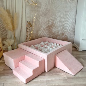 Könnte beinhalten: Rosa Ballgrube mit einer rosa Rampe und Stufen. Die Ballgrube ist mit weißen und rosa Bällen gefüllt. Die Ballgrube befindet sich in einem Raum mit einer weißen Wand und einem hellbraunen Boden.