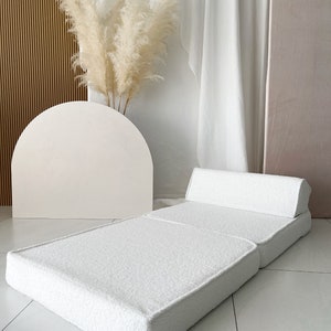 May include: A white, fluffy, foldable futon mattress with a matching pillow. The futon is folded in half and the pillow is placed on top of the folded section. The futon is on a white tiled floor.