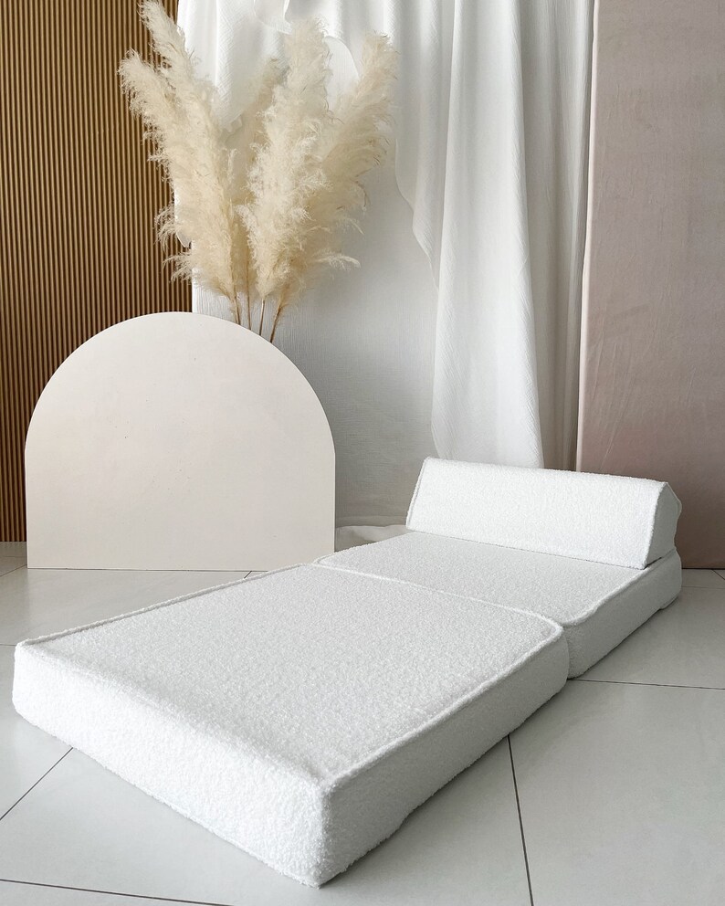May include: A white, fluffy, foldable futon mattress with a matching pillow. The futon is folded in half and the pillow is placed on top of the folded section. The futon is on a white tiled floor.