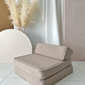 May include: A beige, fluffy, square-shaped floor cushion with a back rest. The cushion is made of a soft, plush fabric and has a simple, modern design.