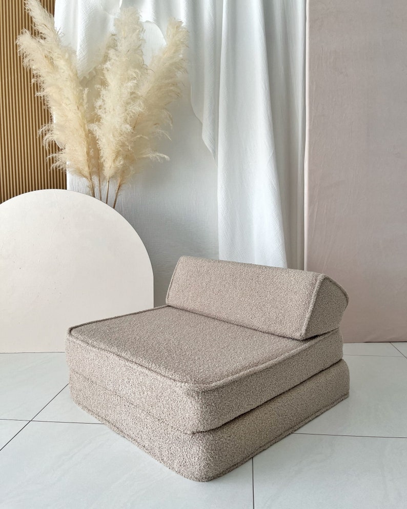 May include: A beige, fluffy, square-shaped floor cushion with a back rest. The cushion is made of a soft, plush fabric and has a simple, modern design.
