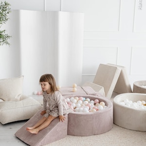 May include: A child sits on a pink corduroy play structure with a ramp, next to a ball pit filled with colorful balls. Other play structures and a ball pit are visible in the background. The scene is set on a light-colored rug.