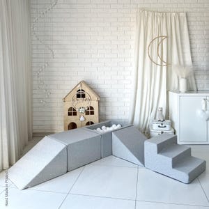 Può includere: Un set di giochi grigio per bambini piccoli, con una piscina di palline, una rampa e una serie di scale. Il set di giochi è realizzato in materiale morbido e imbottito ed è perfetto per incoraggiare il gioco attivo.