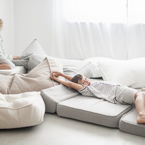 May include: Two children relaxing on a bed of soft, beige and gray cushions. The cushions are arranged in a variety of shapes and sizes, creating a comfortable and inviting space.