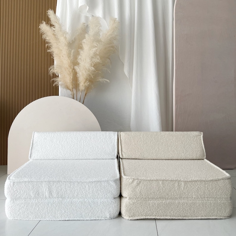 May include: Two white and beige foldable futon chairs with a fluffy texture. The chairs are sitting on a light wood floor in front of a white wall.