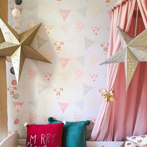 Puede incluir: Una cama con dosel rosa con un papel tapiz estampado en blanco y rosa con una cadena de pompones blancos y dos decoraciones de estrellas doradas. La cama tiene una almohada roja con el texto "Merry Christmas" y una almohada verde.
