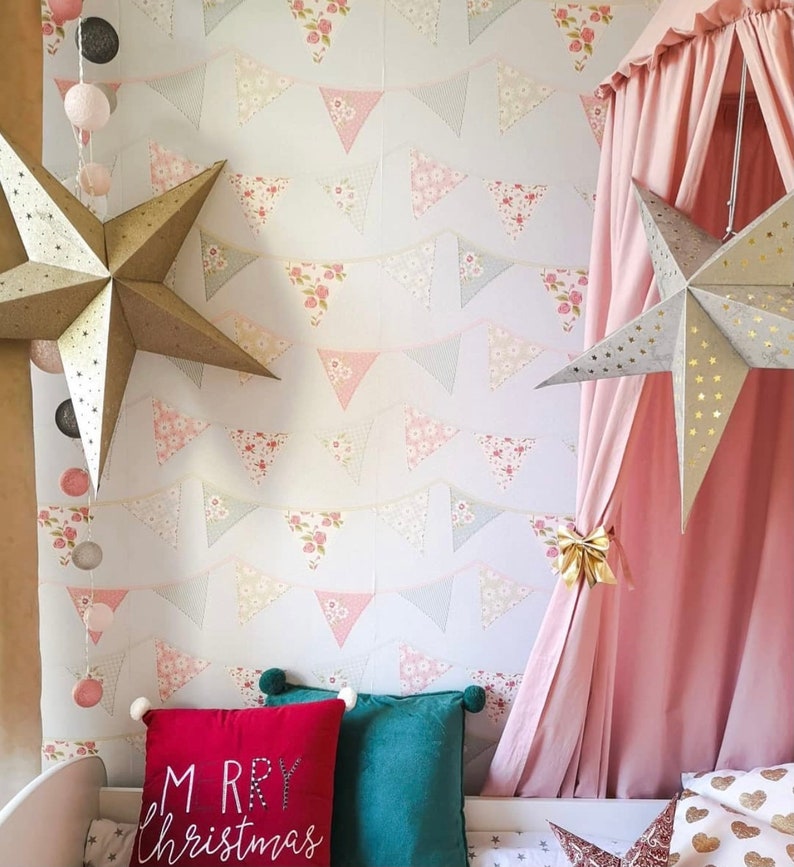 Puede incluir: Una cama con dosel rosa con un papel tapiz estampado en blanco y rosa con una cadena de pompones blancos y dos decoraciones de estrellas doradas. La cama tiene una almohada roja con el texto "Merry Christmas" y una almohada verde.