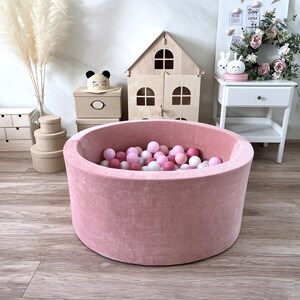May include: A pink velvet ball pit filled with pink and white balls. The ball pit is sitting on a wooden floor in a room with a dollhouse, a nightstand, and a vase of dried flowers.