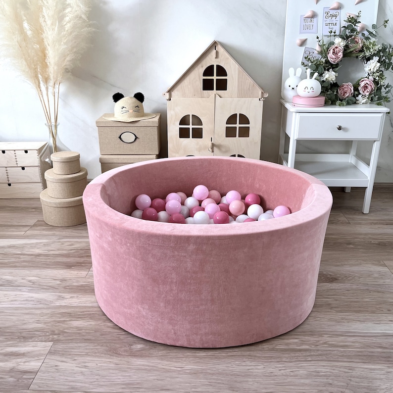 May include: A pink velvet ball pit filled with pink and white balls. The ball pit is sitting on a wooden floor in a room with a dollhouse, a nightstand, and a vase of dried flowers.