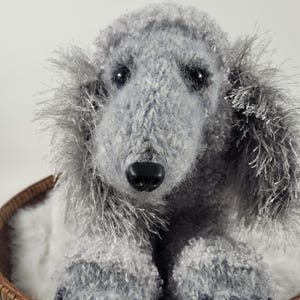 May include: A handmade gray poodle plush toy with a black nose and eyes, resting in a woven brown basket. The poodle has fluffy, textured fur and is positioned facing the viewer, creating a soft and inviting appearance.