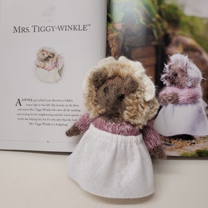 May include: Two plush hedgehog toys wearing pink and white dresses. The hedgehog on the left has a brown and white fur coat and a white bonnet. The hedgehog on the right has a gray fur coat and a white bonnet. The text "MRS. TIGGY-WINKLE™" is visible in the background.