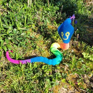 Puede incluir: Una serpiente de crochet arcoíris con una cabeza azul y una cara sonriente. La serpiente está tumbada en un lecho de hierba verde.