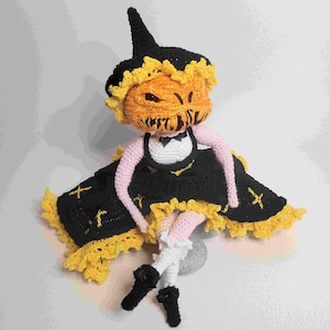 May include: A crocheted pumpkin doll wearing a black and yellow dress with a white lace trim and a black witch hat. The doll has a scary pumpkin face.