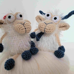 May include: Two white crocheted sheep with blue yarn accents and large googly eyes. The sheep are sitting on a white fluffy surface.
